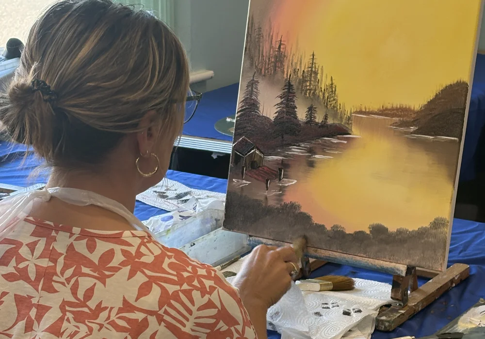 Student painting a golden sunset lake and cabin in Bob Ross wet-on-wet oil at painting class in Kent