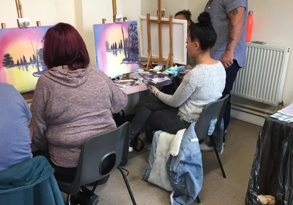 Students painting a Bob Ross sunset lake with silhouette trees at an oil painting class in Kent