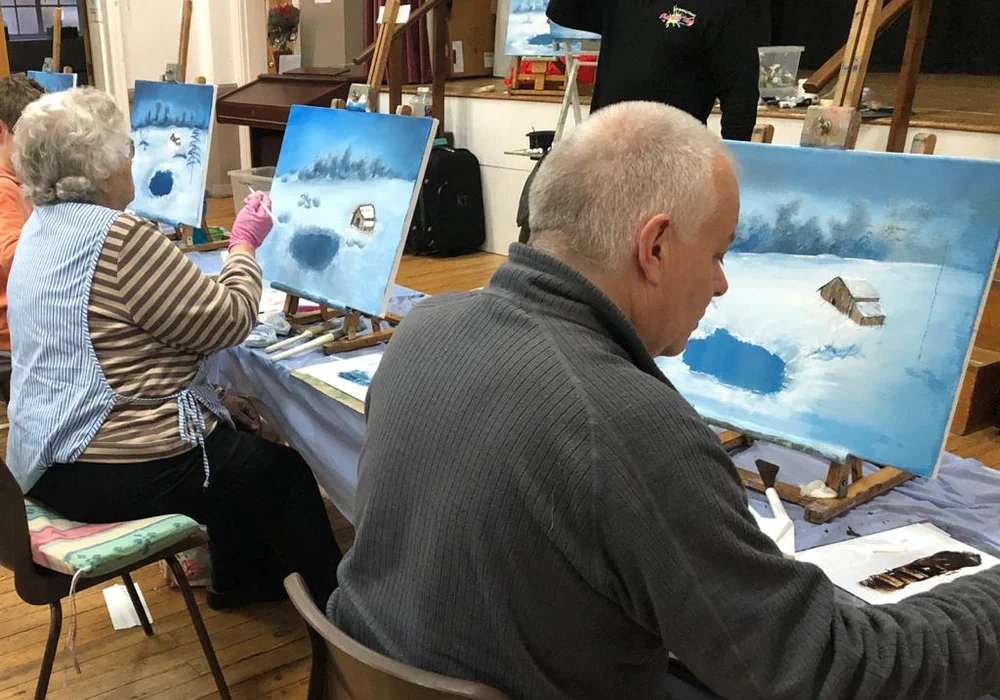 Students painting a Bob Ross winter snow scene with cabin at a Kent oil painting class