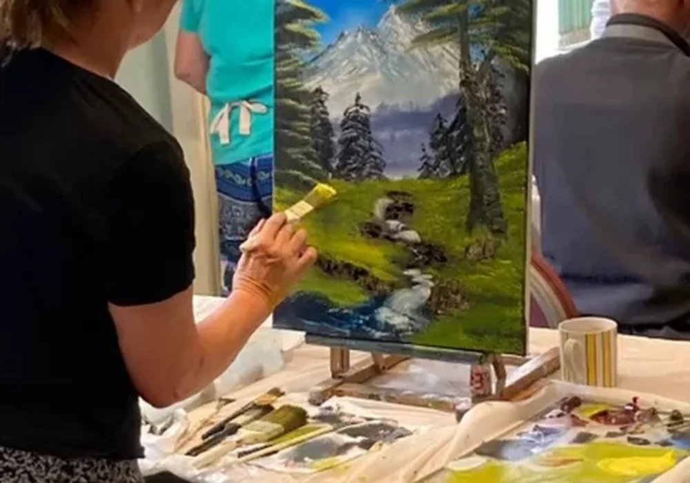 Woman painting a Bob Ross mountain stream landscape at her easel during a Kent painting class